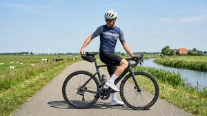 Mannelijke wielrenner met helm en zonnebril zittend op zijn racefiets op een zonnige Nederlandse polderweg, terwijl hij achterom reikt naar zijn zadeltasje.