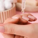 Woman applying oil onto cuticles on pink background, closeup