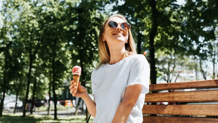 Joyful young woman relaxing on a park bench looking up enjoying the sun and summer weather. Cute smiling girl eating ice cream in a waffle cone, outdoors. Summer lifestyle