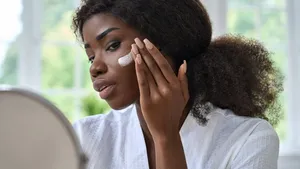 Attractive black young woman applying cream on face skin looking in mirror.