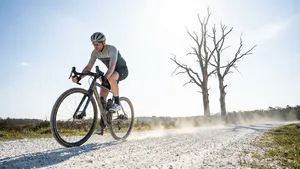 Mannelijke wielrenner op gravelbike op verticale foto, rijdend op brede witte grindweg. Helm en voorjaarskleding, met de kenmerkende dode bomen op de achtergrond.
