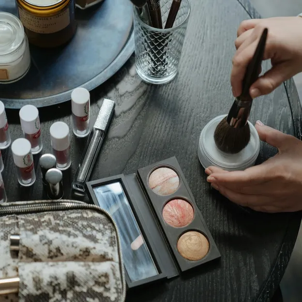 Zo lang blijft make-up houdbaar – Top-down foto van make-up op een tafel. Twee handen houden een doosje en een kwast vast.