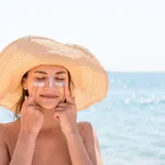 Beautiful young woman in hat is applying sunblock under her eyes and on her nose like Indian. Sun protection concept