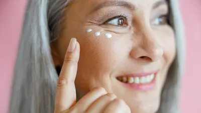 Close-up van een vrouw die anti-aging oogcrème onder haar ogen aanbrengt, in een studio tegen een roze achtergrond.