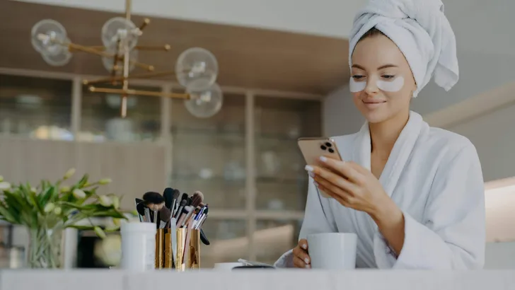 Photo of attractive young woman wears bath towel on head and soft white dressing gown holds mobile phone enjoys online communication applies moisturising undereye patches drinks hot beverage