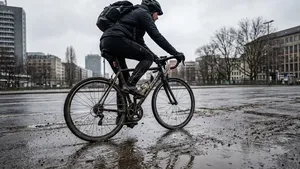 Actiefoto van een fietser op een racefiets, van schuin achteren gezien, die op een nat fietspad rijdt terwijl water en vuil opspatten van het achterwiel.