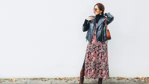 Smiling female dressed boho fashion style colorful long dress with black leather biker jacket with brown leather flap bag posing on the white wall background.