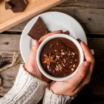 Girl drinks hot chocolate mug, with christmas present on rustic table with blanket or plaid from above, cozy and tasty breakfast or snack. Hands in picture, top view