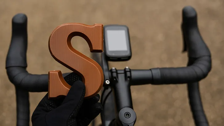Close-up van een wielrennerhandschoen die een melkchocoladeletter S vasthoudt voor het stuur van een racefiets, met het fietsstuur en de fietscomputer onscherp op de achtergrond.