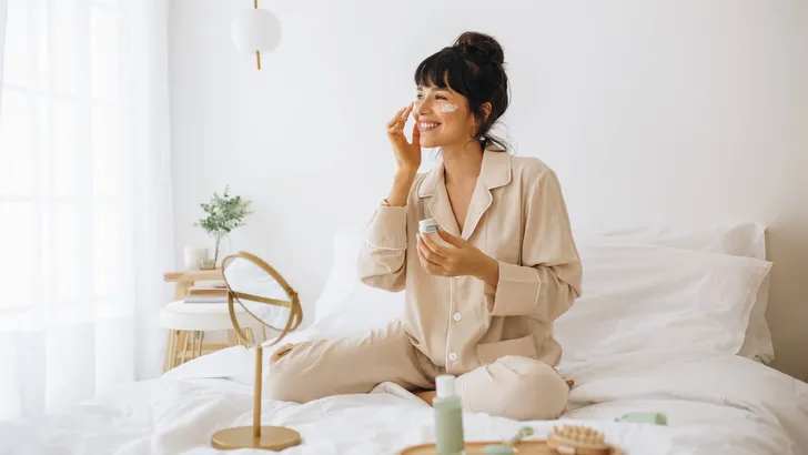 Smiling woman applying face cream sitting on bed