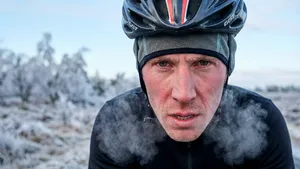 Een wielrenner met helm en muts kijkt intens in de camera, met zichtbare ademwolkjes door de vrieskou in een bevroren landschap.