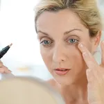 Middle-aged blond woman putting eye concealer in front of mirror