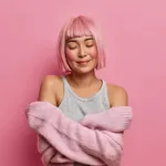 Women, tenderness, comfort. Pleased pink haired Asian woman embraces herself, closes eyes, feels coziness in soft sweater, stands against rosy background, dreams about something in peace atmosphere