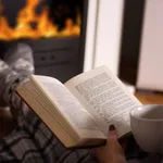 Woman reading book and hold cup of tea and warming feet in front of the fireplace