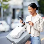 Happy woman with her phone, credit card and bag after shopping in the city. Young latin female carrying bags, spending money, looking for sales and enjoying online eCommerce store sale with a smile