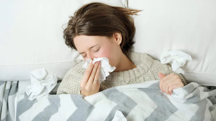 A young woman lies in bed under a blanket and blows her nose in