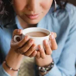 Close up of young beautiful unrecognizable woman hands holding hot cup of coffee or tea with milk. Morning coffee, office coffee break or coffee lover concept.