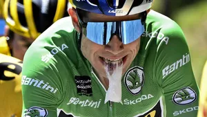 Close-up van Wout van Aert in groene trui die tijdens de koers een gel inneemt, met geel-zwarte ploegen op de achtergrond.