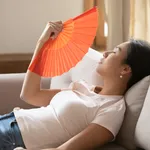 Tired young asian woman using paper fan indoors.