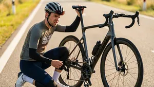 Mannelijke wielrenner in Wieler Revue-kleding met arm- en beenstukken en een helm knielt langs een zonnige weg en controleert de onderdelen van zijn racefiets op een tikkend geluid.