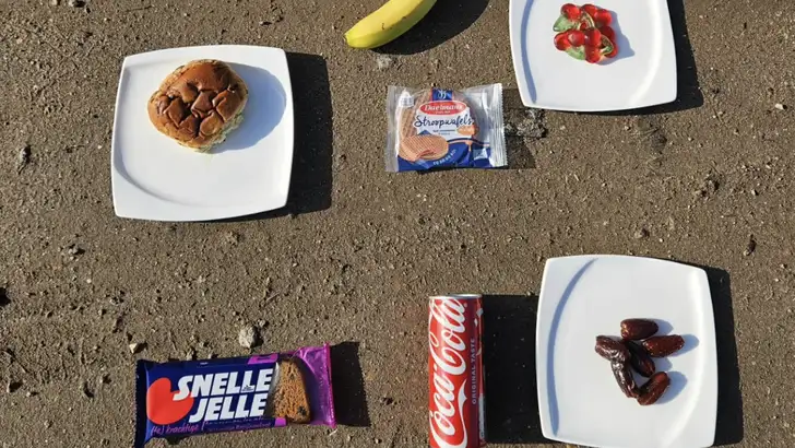 Overzicht van diverse fietssnacks, waaronder een Mars, banaan, stroopwafel, Snelle Jelle, Coca-Cola blikje, dadels, Haribo Cherries en een krentenbol, liggend op een zanderige ondergrond.