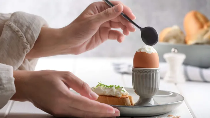 Woman eating fresh soft boiled egg at white wooden table, closeup