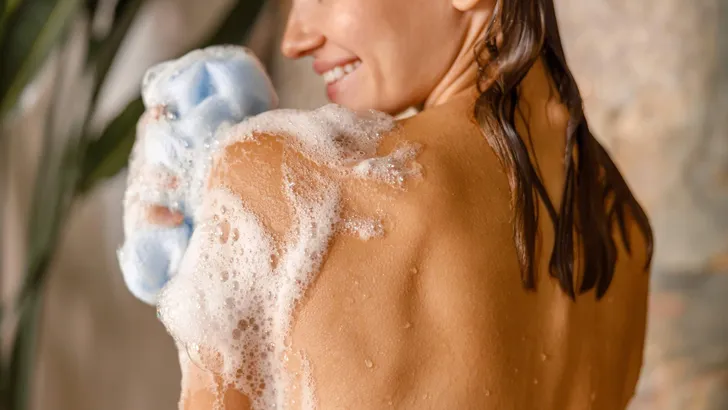 Happy young woman applying shower gel on her body using loofah sponge while taking shower