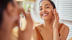 Smiling mixed race young woman applying moisturizer on her face in bathroom