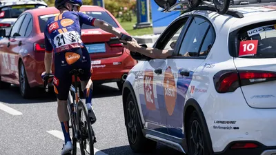 en profwielrenner van Team Picnic-PostNL houdt stevig een bidon vast die wordt aangereikt vanuit het raam van de ploegleidersauto, het klassieke beeld van een plakbidon.