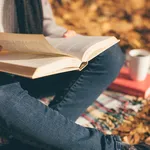 Cropped image of young woman sitting on blanket, reading book and drinking coffee or tea in autumn garden