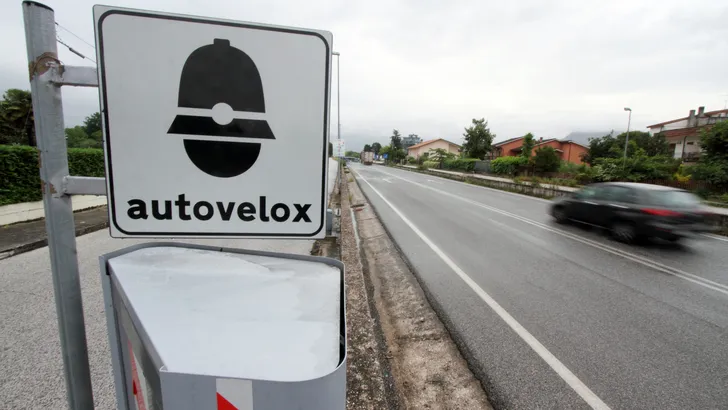 Italiaans autovelox-bord en flitspaal langs een natte weg, met een voorbijrijdende vrachtwagen op de achtergrond.