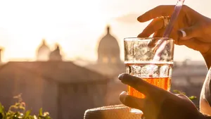 Young woman tourist fashion white dress with spritz cocktail in front of panoramic view of Rome cityscape from campidoglio terrace at sunset. Landmarks, domes of Rome, Italy.