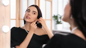 Young woman trying on elegant pearl earring near mirror indoors