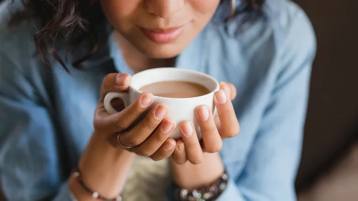 Close up of young beautiful unrecognizable woman hands holding hot cup of coffee or tea with milk. Morning coffee, office coffee break or coffee lover concept.