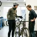 Een wielrenner in winterkleding brengt zijn racefiets voor een onderhoudsbeurt naar een fietsenmaker, met een kalender die 'FEBRUARI' aangeeft op de achtergrond.