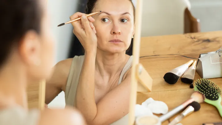 Een vrouw van middelbare leeftijd kijkt in kleine spiegel en gebruikt make-up om haar wenkbrauwen voller te maken.