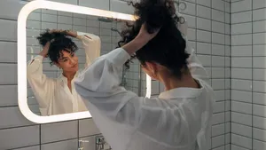 Young mixed race woman looking herself in the mirror in modern washroom. Girl makes a bun in her curly hair. Female making herself a ponytail.