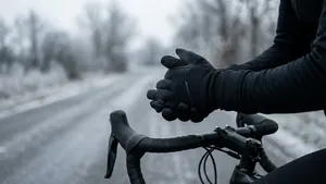 Een wielrenner op een racefiets wrijft in zijn handen om ze warm te houden, draagt zwarte winter fietshandschoenen op een koude, grijze winterdag met een berijpte weg op de achtergrond.