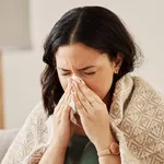 Nose, tissue and sick woman sneezing on a sofa with allergy, cold or flu in her home. Hay fever, allergy and female with viral infection, problem or health crisis in a living room with congestion