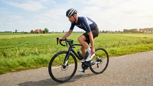 Een mannelijke fietser in blauw-witte wielerkleding en een helm rijdt op een racefiets over een asfaltweg door een vlak Nederlands polderlandschap met molens in de verte en grasvelden onder een deels bewolkte lucht.