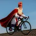 Sinterklaas rijdt op een racefiets over dakpannen onder een heldere blauwe lucht, met een wapperende rode mantel voor een humoristisch Sinterklaas-en-wielrennenbeeld.