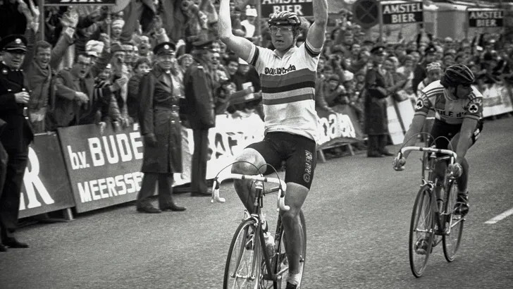 Jan Raas juicht met zijn armen in de lucht terwijl hij in de iconische regenboogtrui de Amstel Gold Race wint.