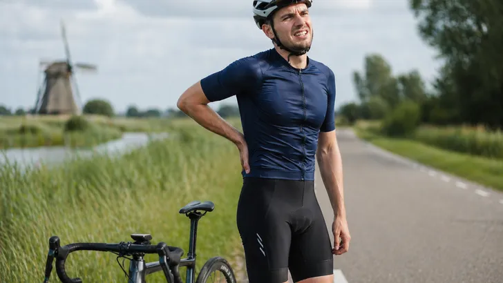 Mannelijke wielrenner staat naast zijn racefiets op een rustige Nederlandse landweg, met één hand op zijn onderrug en een licht pijnlijke gezichtsuitdrukking