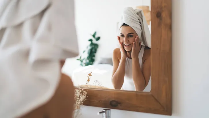 Young beautiful female with towel on head smiling to mirror refl