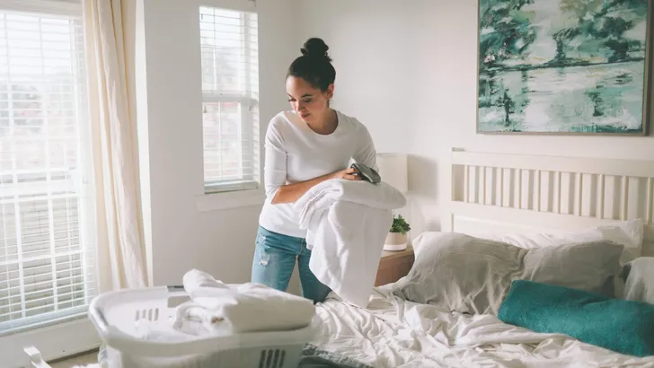 A woman folding laundry in a bright white bedroom.