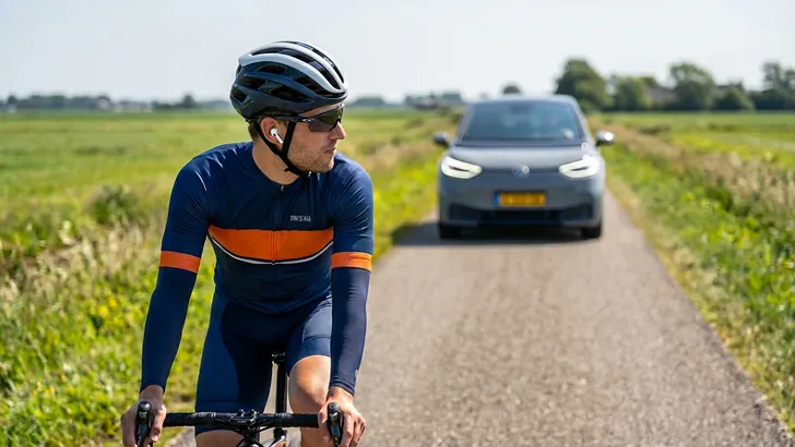 Wielrenner met in-ear oortje fietst solo op een polderweg, met de tekst "Hoor jij het gevaar?" en een naderende auto op de achtergrond.