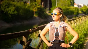 Vrouw in sportkleding en met een hardloopvest met drinkreservoir staat op een landweg op een zonnige dag.