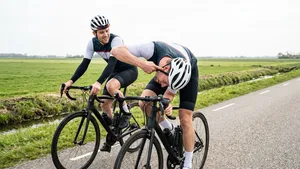 Een paranoïde wielrenner met helm buigt zich tijdens het fietsen vreemd naar beneden om naar een kraakje te luisteren, Gadegeslagen door een verbaasde fietsmaat.