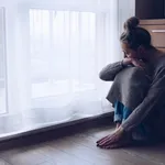 A devastated sad woman is sitting on the floor in her living room looking at the window.