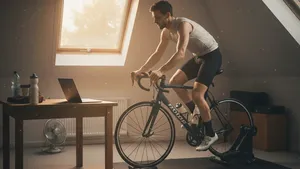 Wielrenner met helm en korte fietskleding staat op de pedalen van een indoorfietstrainer op een zolderkamer, terwijl hij naar een laptop kijkt die op een tafel voor hem staat.